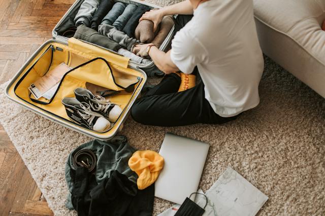 person-packing-clothes-in-a-suitcase-with-clothes-a-laptop-and-shoes-around them