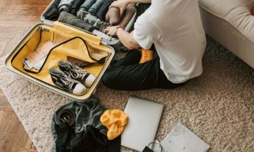 person-packing-clothes-in-a-suitcase-with-clothes-a-laptop-and-shoes-around them